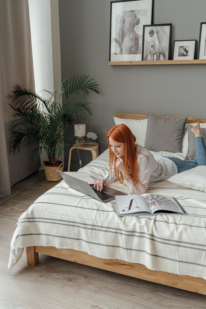 Red-haired woman working remotely on a laptop in a contemporary bedroom setting.